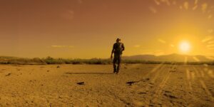 Seen from behind, a man moves on foot across dried and cracking ground; the sun blazes on the horizon, at the right (iStock)